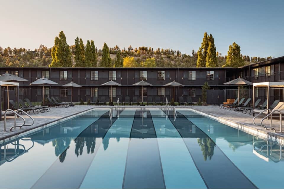Olympic-size outdoor swimming pool with lounge chairs, modern resort building, and pine forest backdrop