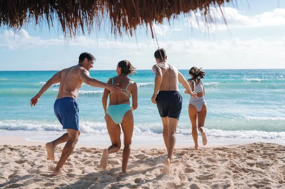 Family running into turquoise ocean waves on tropical beach under thatched shelter