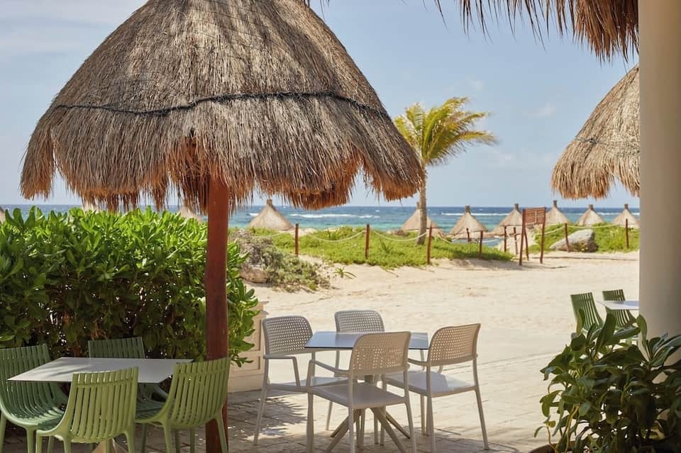 Beachfront dining area under thatched umbrella with ocean view, sand, beach vegetation, and multiple cabanas