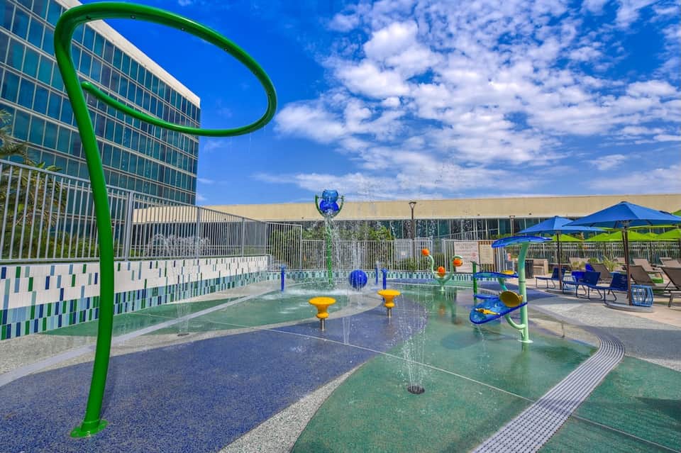 Colorful splash pad at hotel pool area with blue umbrellas, water features, and modern building backdrop