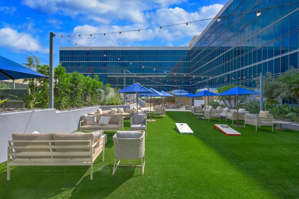 Rooftop terrace with cream-colored lounge furniture, blue umbrellas, artificial grass, and modern glass building overlooking cityscape