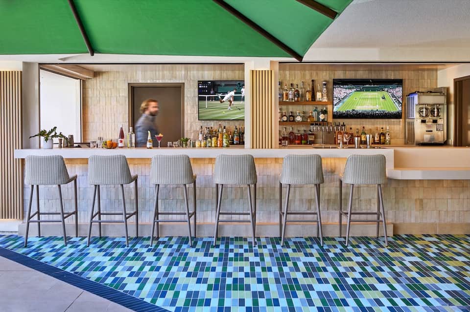 Modern hotel bar with six bar stools, shelved liquor bottles, and green ceiling detail overhead