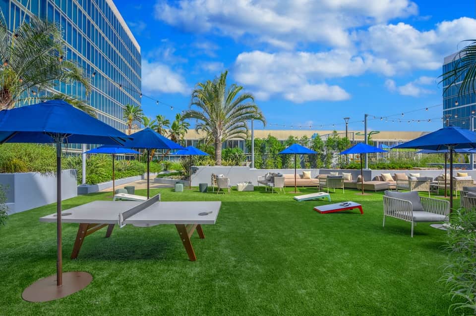 Lawn games area with cornhole boards, blue umbrellas, seating, and palm trees under blue sky