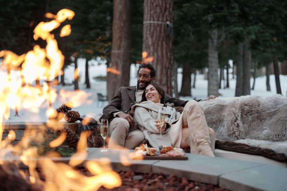 Couple by firepit at resort during evening with fire, blankets, drinks, and forest backdrop
