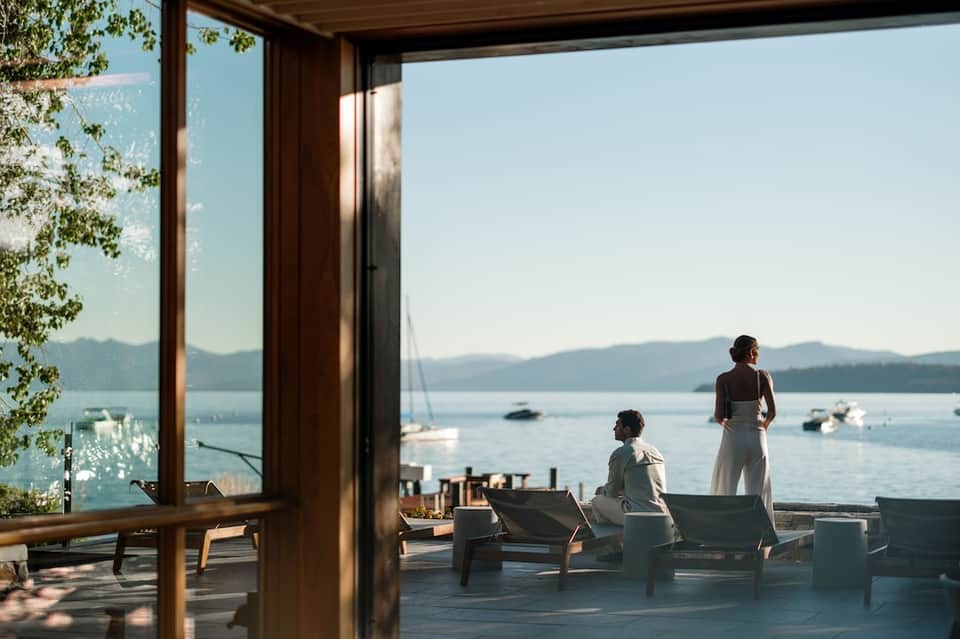 Two people relaxing on lounge chairs on a lakeside terrace with water views, boats, and mountain scenery visible