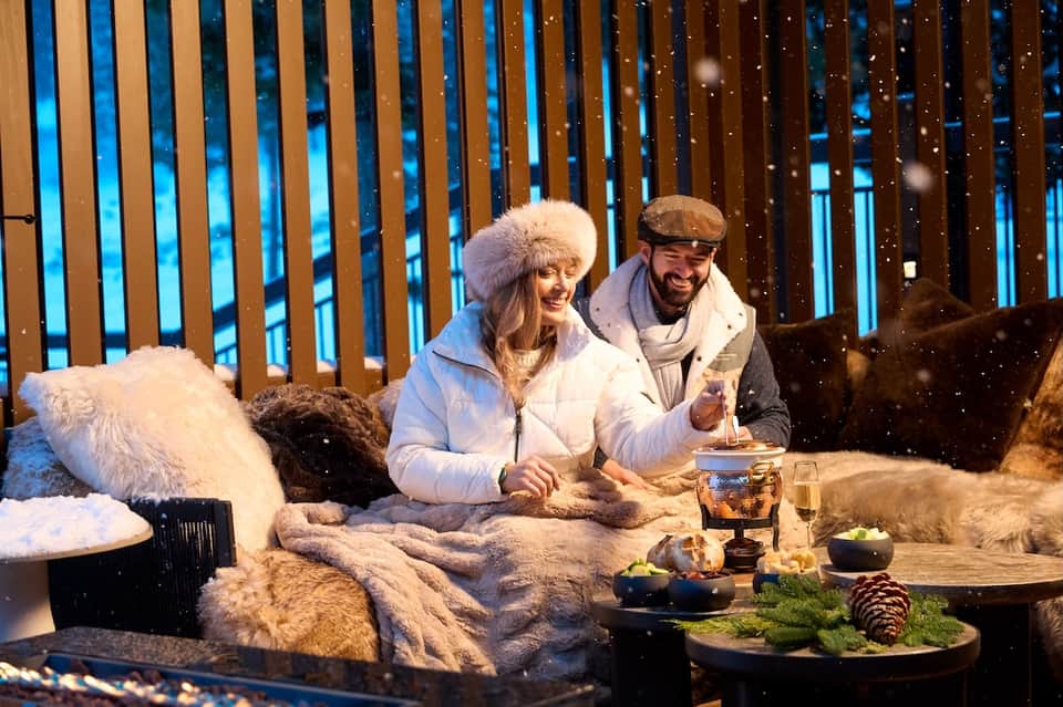 Couple enjoying drinks on snowy balcony overlooking nighttime ski resort and mountains