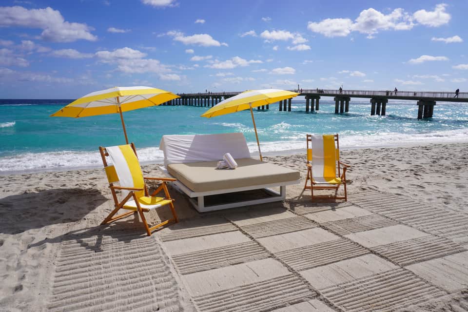 Beach lounge setup with yellow umbrellas, cushioned daybed, and wooden pier extending into turquoise ocean