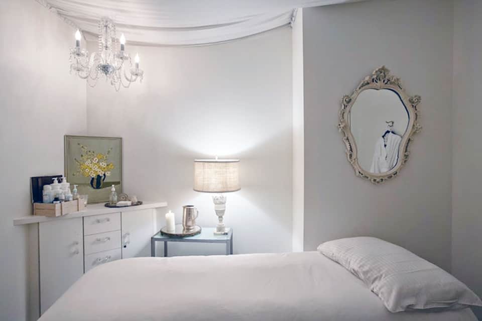 Minimalist spa bedroom with white bedding, grey walls, ornate mirror, and soft table lamp