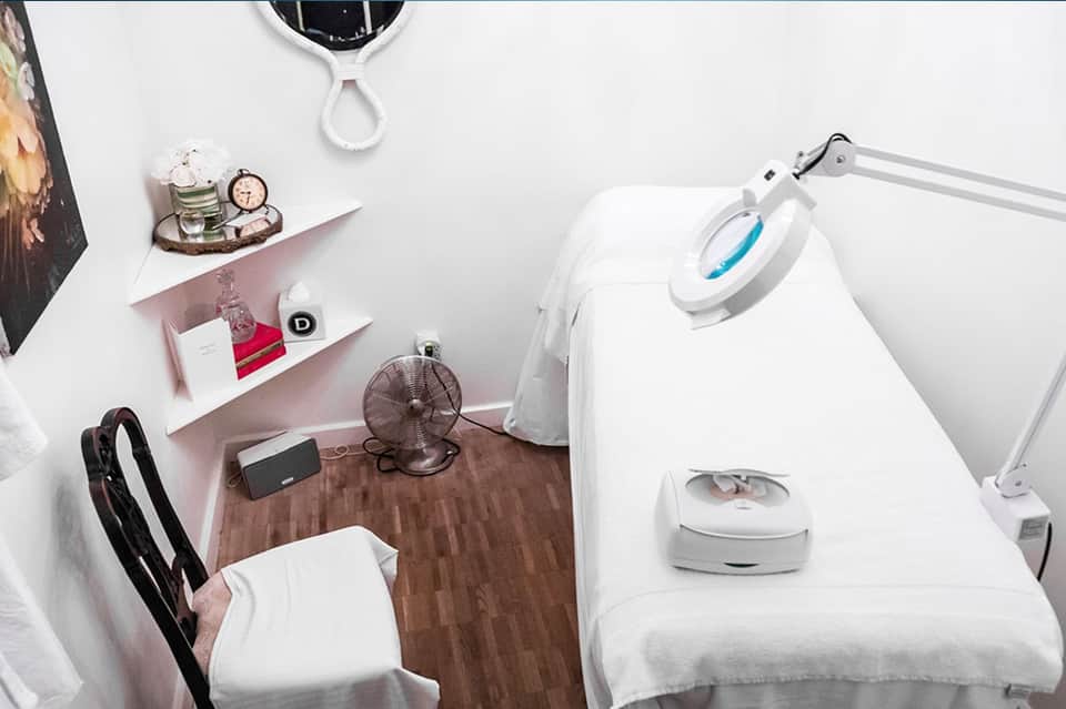 Modern spa treatment room with white bed, professional equipment, shelving, and wood flooring