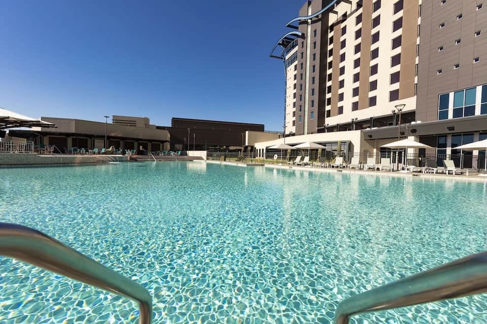 Large resort pool with crystalline turquoise water, tall hotel building, white umbrellas, and clear sunny conditions