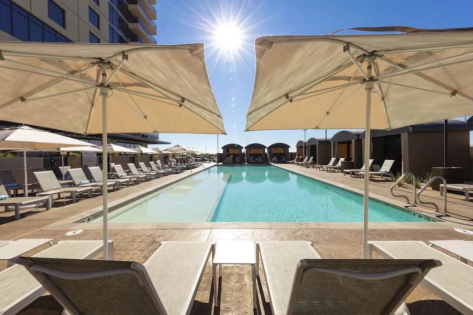 Rectangular pool with large tan umbrellas and loungers, with dome cabanas visible in background under bright sun
