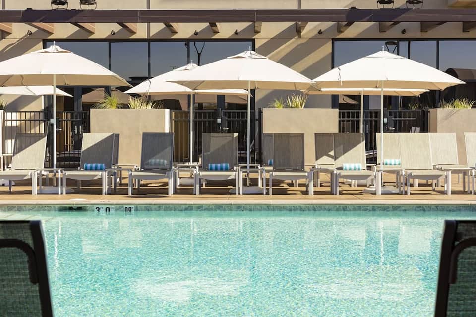 Pool deck with white umbrellas, cushioned loungers, and modern building featuring tall windows and pergola structure