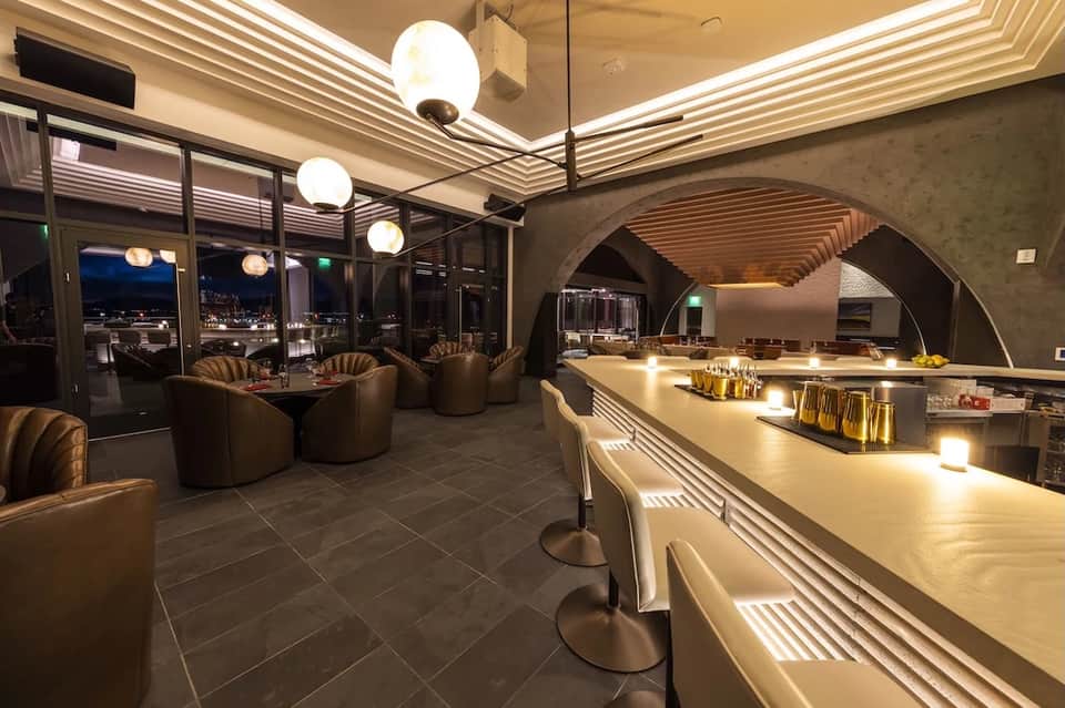Modern lounge bar interior at night with ambient lighting, curved seating, and city views through floor-to-ceiling windows
