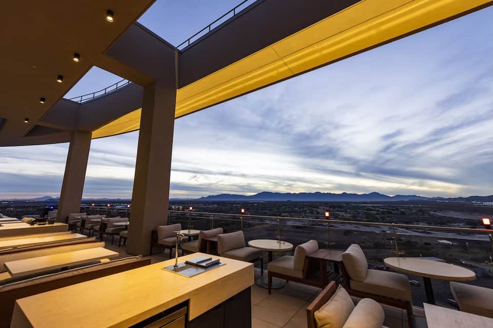 Elevated rooftop lounge at dusk with yellow lighting, modern furniture, and panoramic mountain landscape views