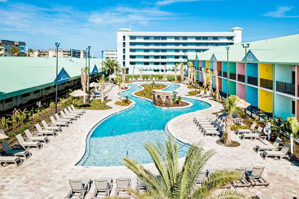 Resort pool area with colorful cabanas, curved blue water features, palm trees, and modern hotel building in background