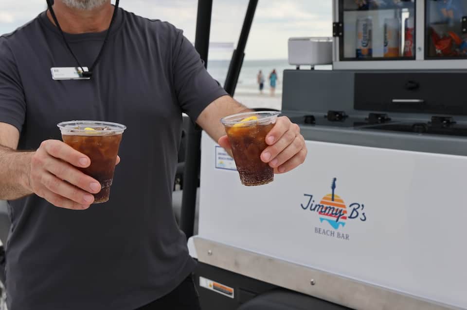 Beach bar server holding two cola drinks with ocean and beachgoers in background