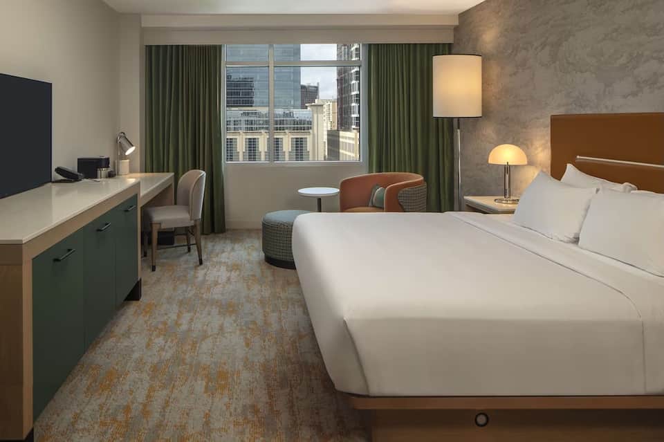 Modern hotel bedroom with king bed, work desk, green curtains, and city window views