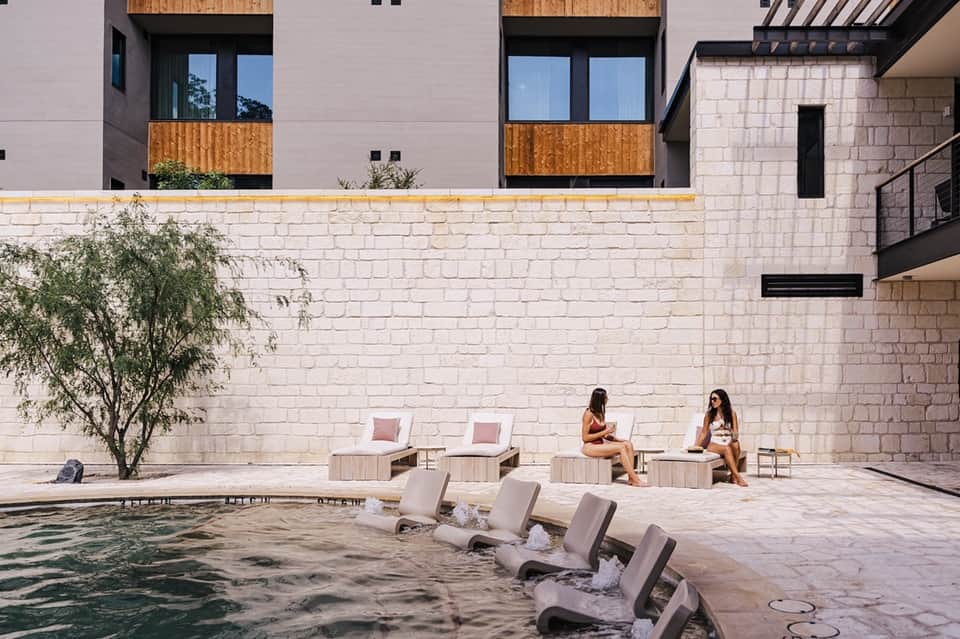 Two women relaxing by courtyard pool with concrete loungers and stone building walls