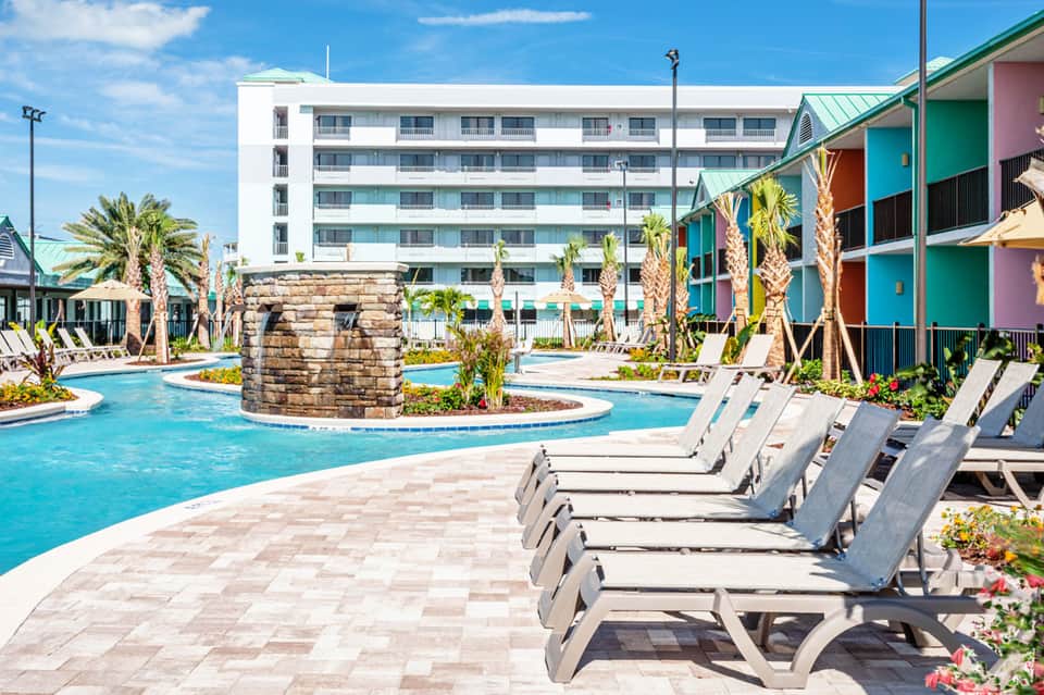 Resort pool area with colorful cabanas, loungers, and multi-story hotel building under blue sky