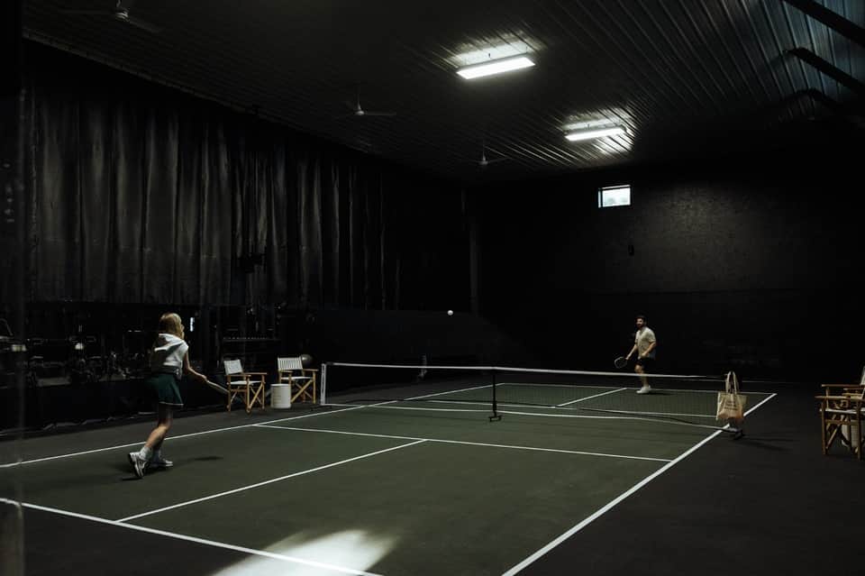 Indoor pickleball court with players, fluorescent lighting, and spectator seating