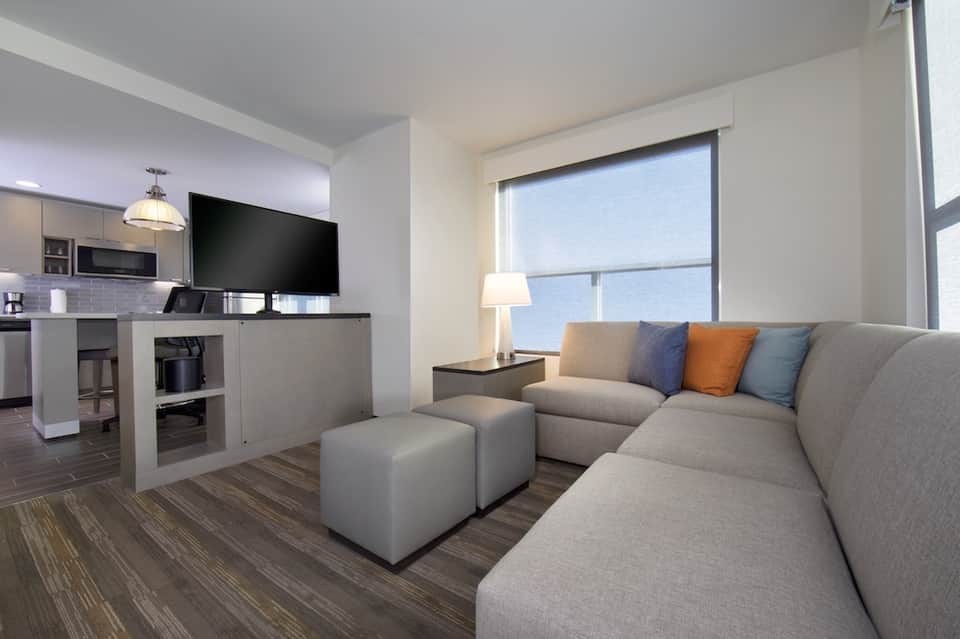 Modern hotel suite with gray sofa, kitchen area, TV, and large bright windows