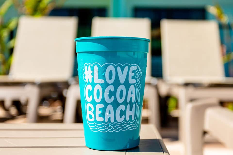 Turquoise reusable cup with #LoveCocoaBeach text on a pool deck with lounge chairs