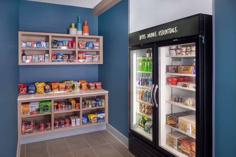 Hotel snack station with wooden shelving, vending refrigerator, and colorful packaged snacks and beverages.