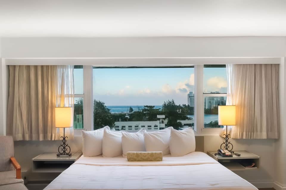 Hotel bedroom with ocean view windows, white bedding, elegant lamps, and coastal scenery