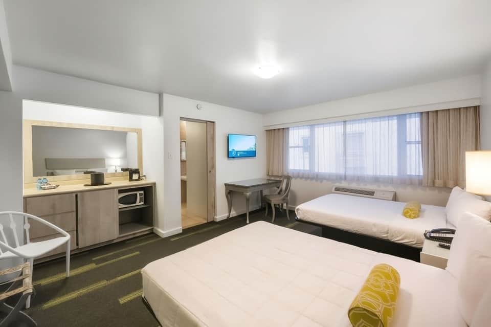 Modern hotel room with two beds, kitchenette with microwave, desk, window with curtains, and minimal decor