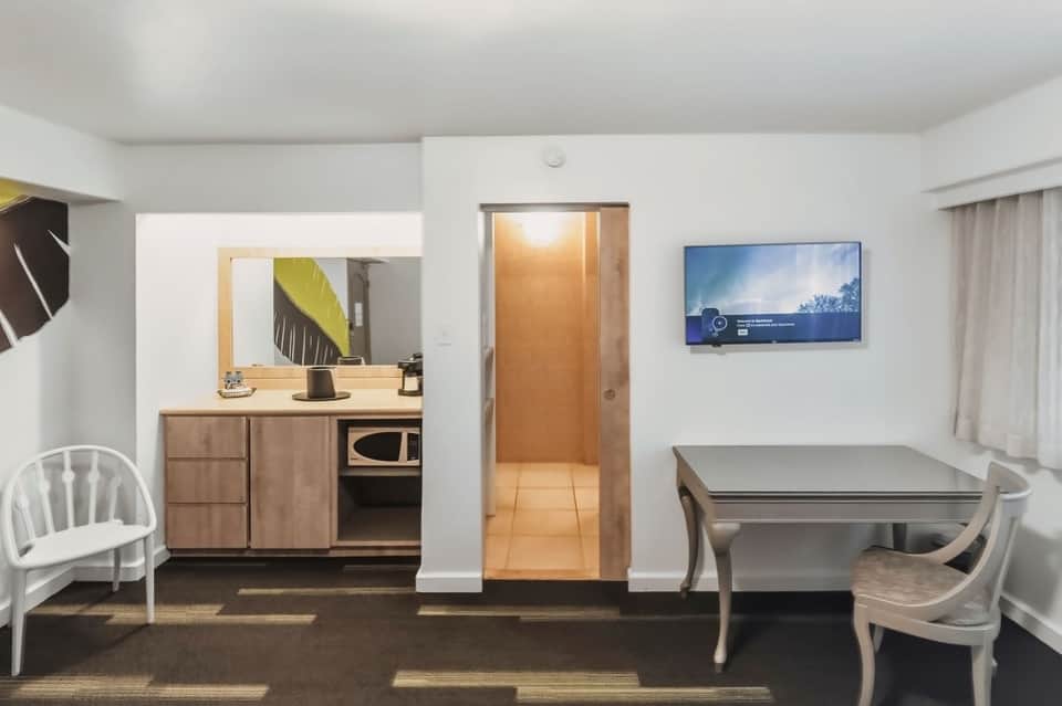 Hotel room kitchenette and living area with wood cabinetry and glass dining table