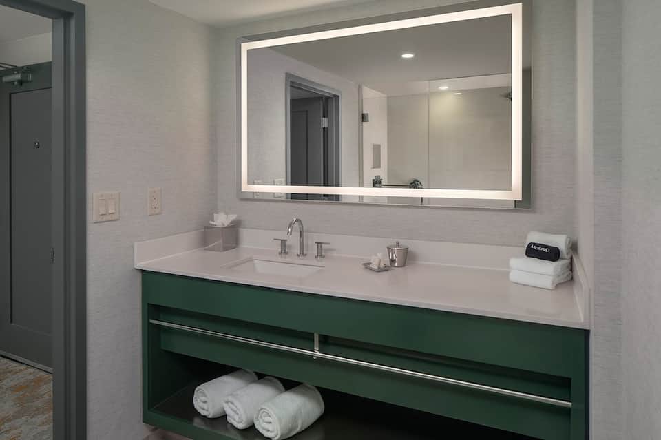 Modern hotel bathroom with illuminated mirror, green vanity cabinet, white countertop, and folded towels