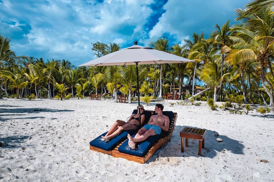 Couple relaxing on blue chaise lounges under large white umbrella on white sand beach with palm trees and clear sky