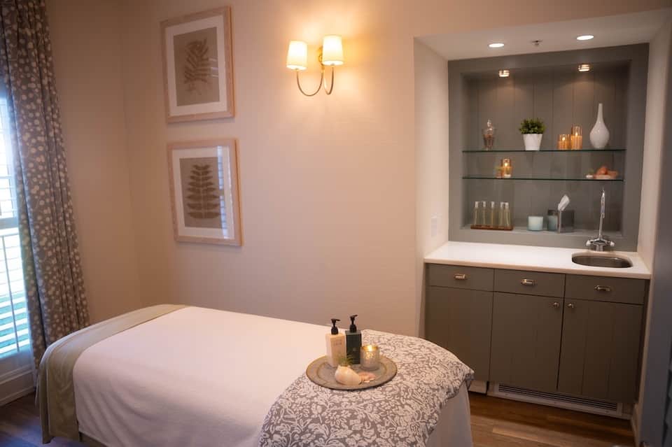 Luxury spa treatment room with massage table, ambient lighting, decorative shelving, and soft furnishings.