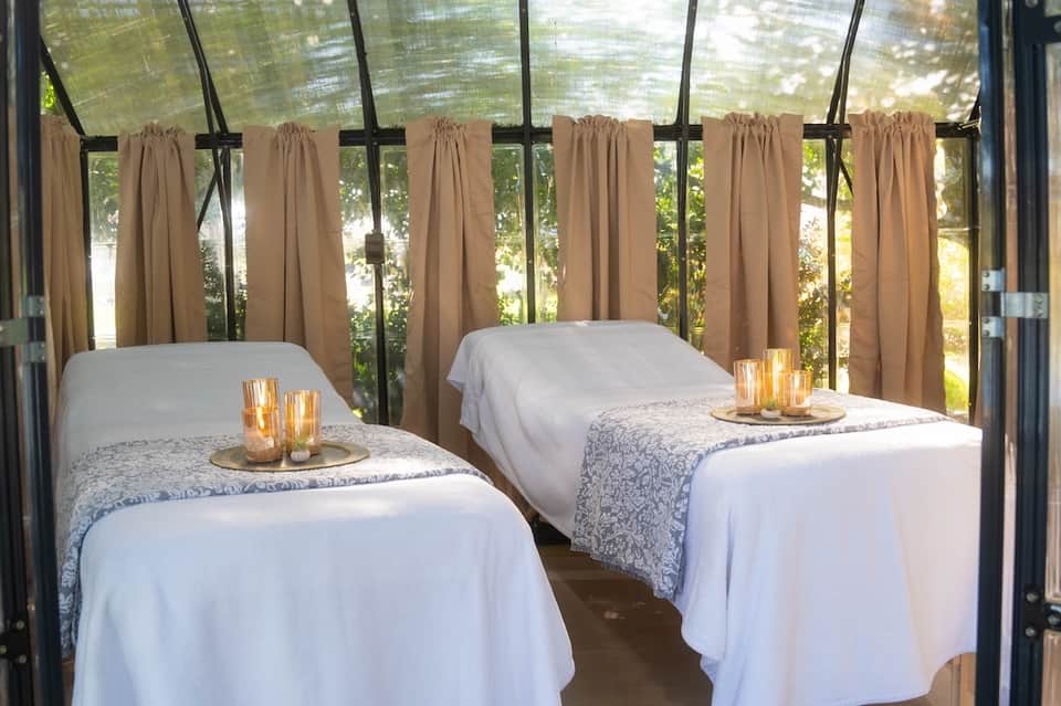 Spa treatment room with two massage beds, sheer curtains, candlelit ambiance, and garden views