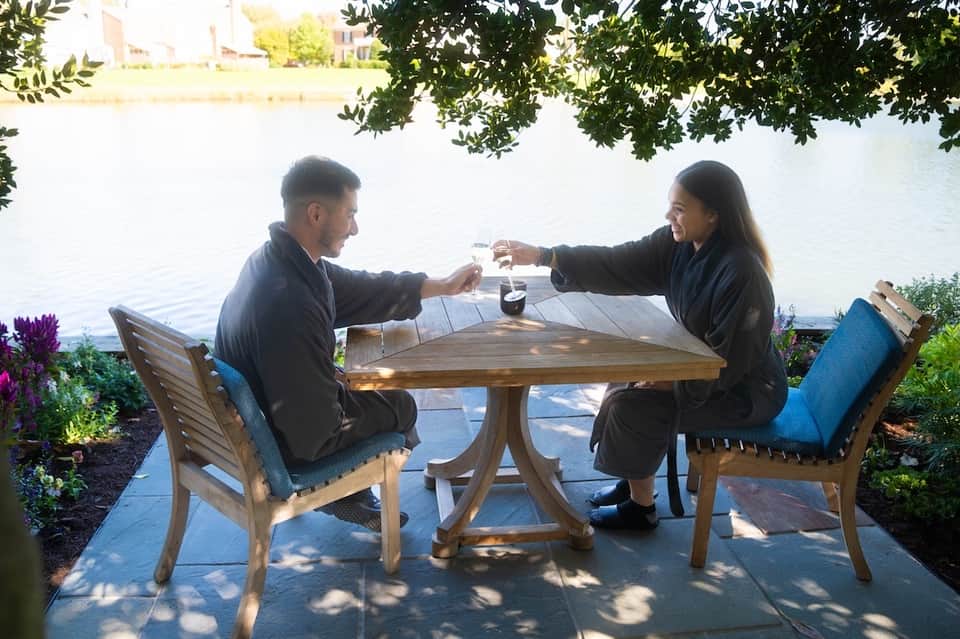 Couple toasting drinks at waterside table with tree shade and scenic water backdrop