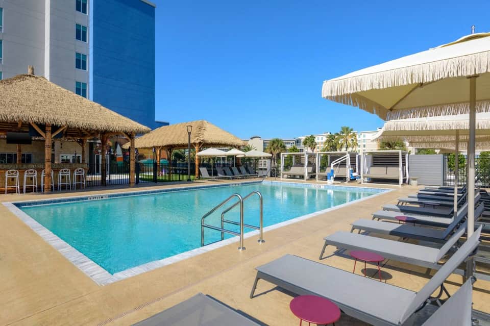 Resort pool deck with thatched-roof bar, lounge chairs, umbrellas, and modern high-rise hotel building