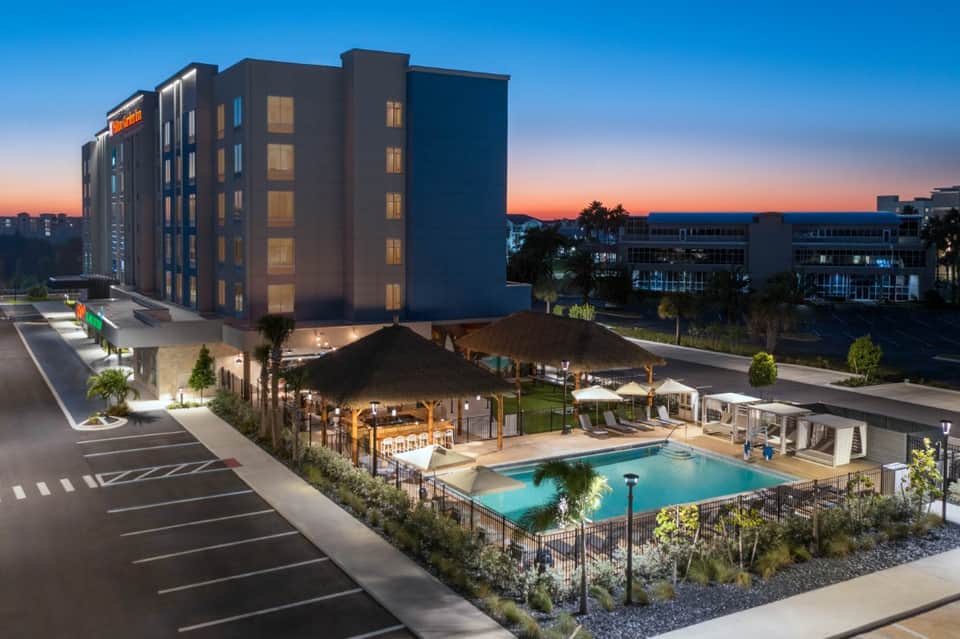 Modern hotel at dusk with illuminated rooms, ground-level pool, covered patio area, and palm trees