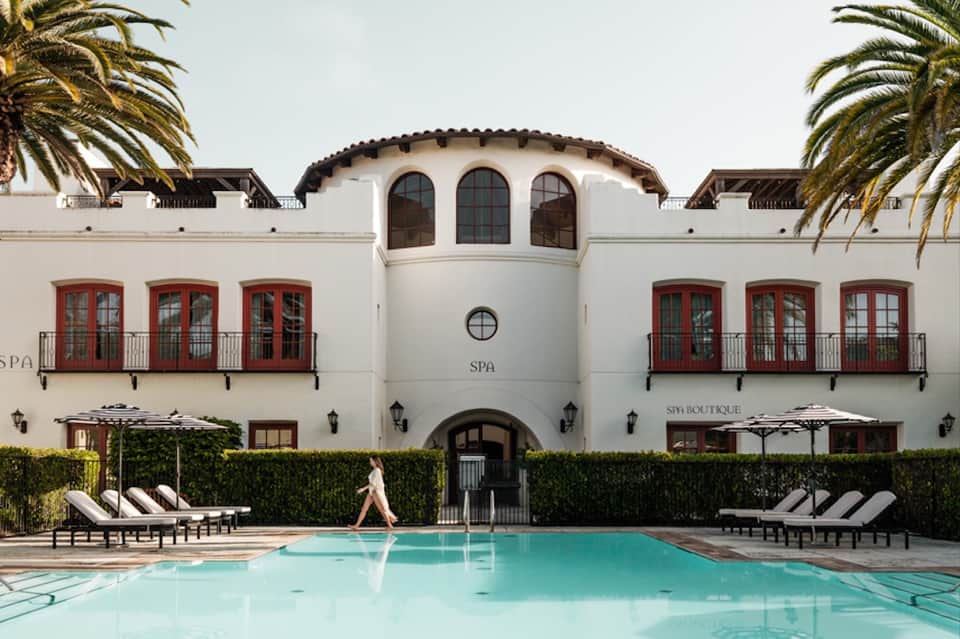 Elegant spa building with white facade, red doors, palm trees, and turquoise pool with loungers