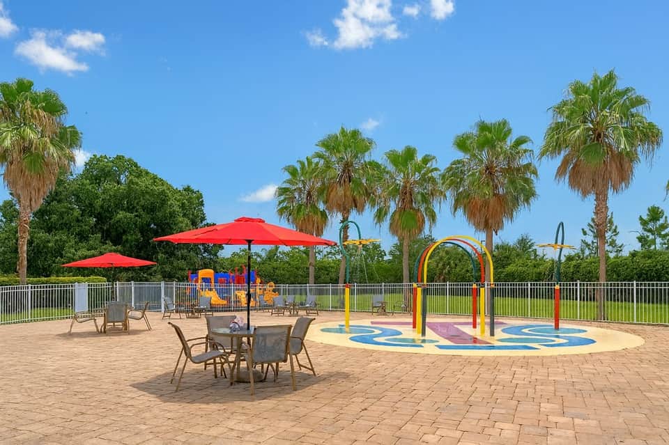 Outdoor playground with red umbrellas, colorful splash pad, and palm trees
