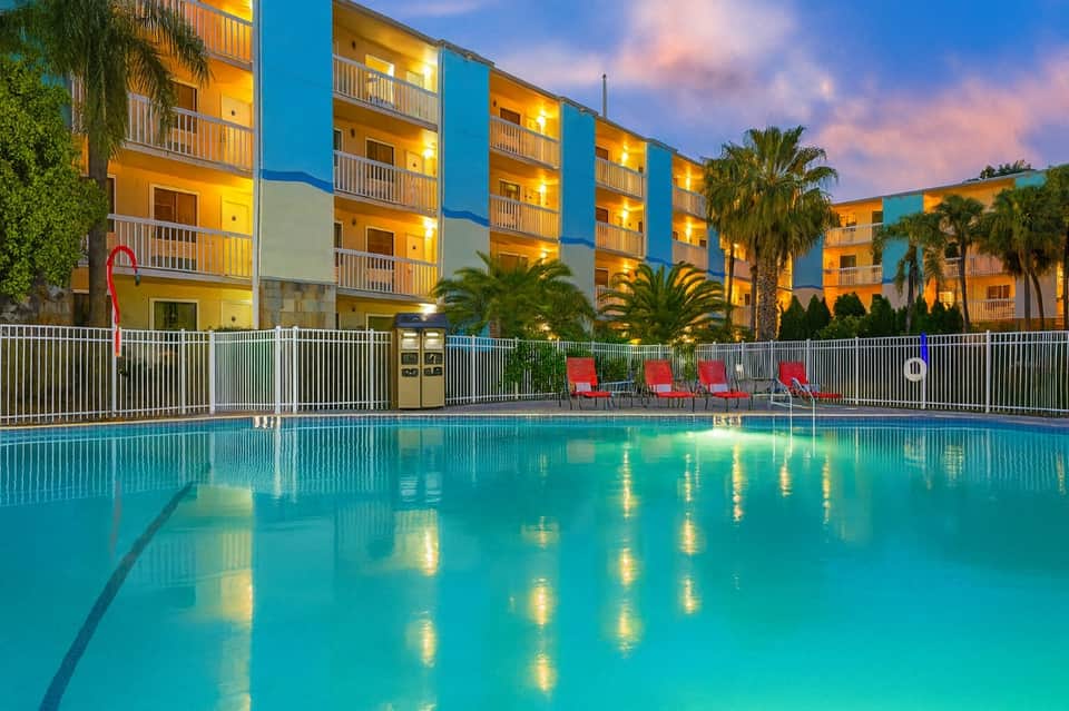 Illuminated hotel building reflected in glowing pool at dusk with red lounge chairs and palm trees