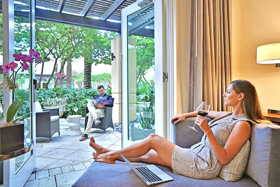 Woman relaxing on sofa with laptop, viewing tropical courtyard garden through floor-to-ceiling windows