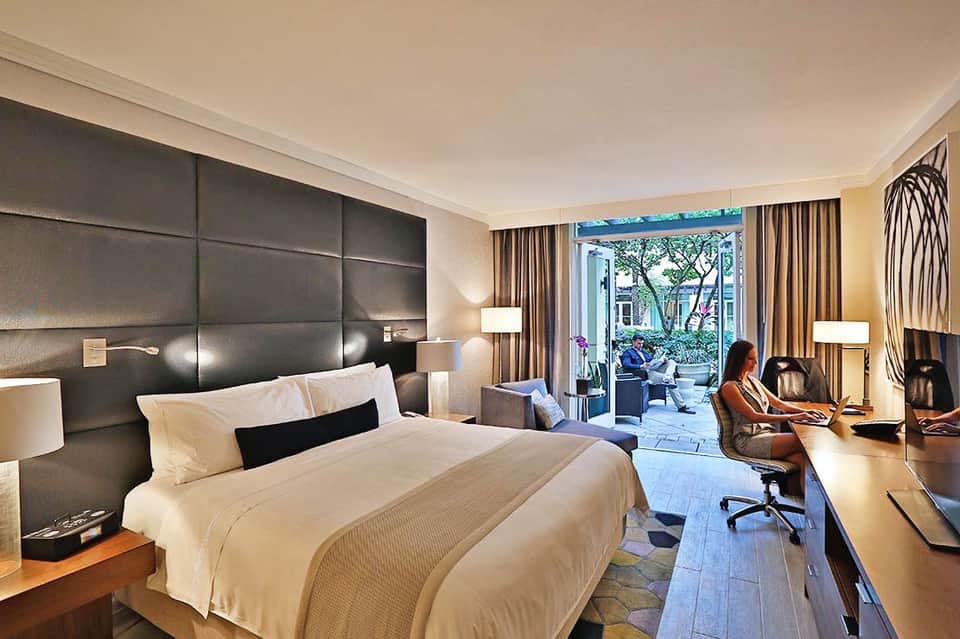 Modern hotel bedroom with dark headboard, king bed, work desk, and balcony access to courtyard