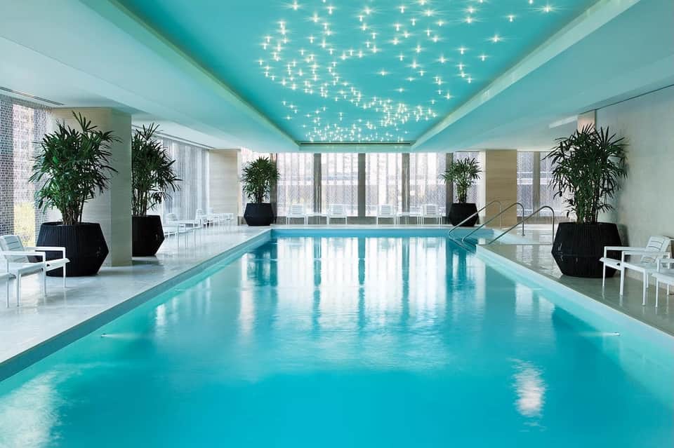 Modern indoor swimming pool with turquoise water, star-lit ceiling, potted plants, and floor-to-ceiling windows