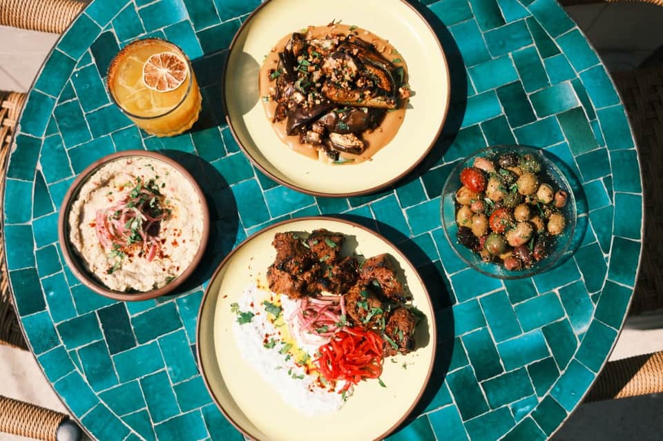 Mediterranean-style appetizers on turquoise tiled table: hummus, grilled vegetables, bread, and fresh juice