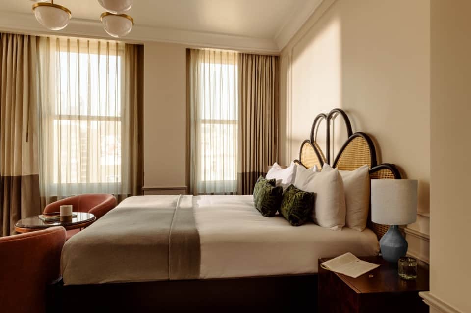 Upscale bedroom with rattan headboard, white bedding, green accents, and large draped windows