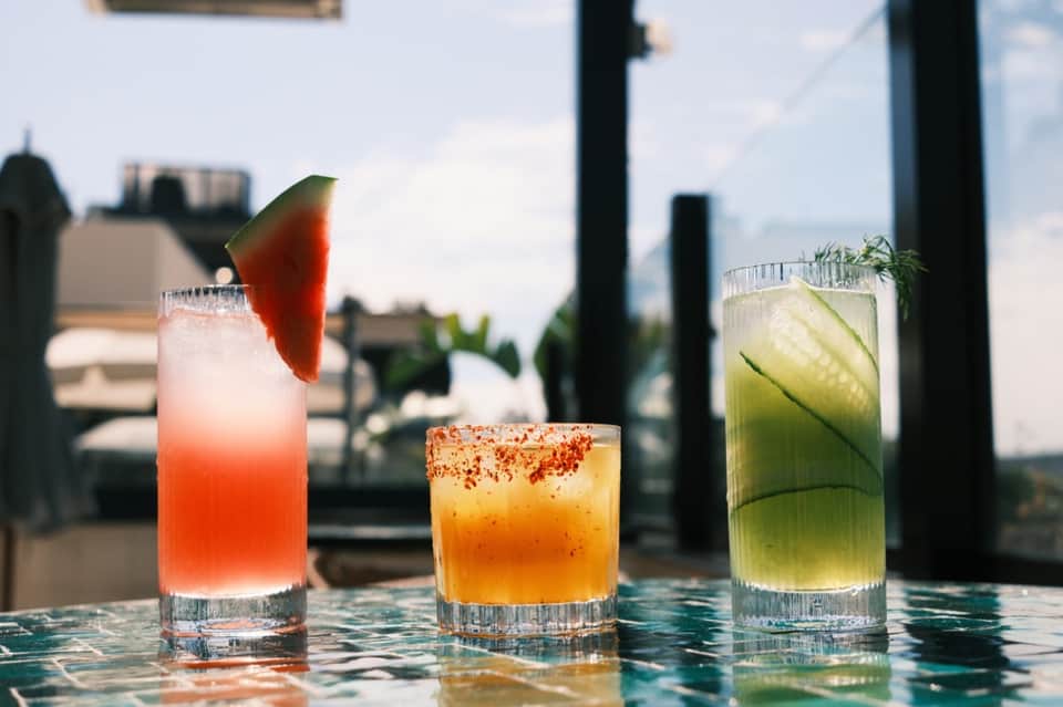 Three colorful cocktails on outdoor bar table with rooftop city views in background