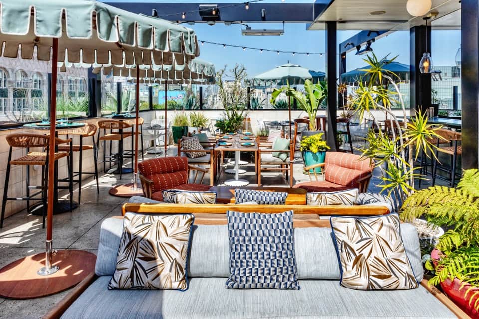 Rooftop restaurant terrace with colorful seating, dining tables, mountain views, plants, and string lights