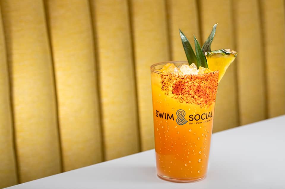 Signature cocktail with Tajín rim, pineapple garnish, and Swim & Social branding on yellow curtain background