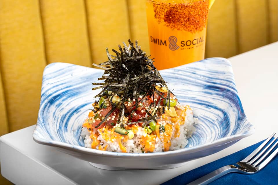 Gourmet sushi bowl with crispy nori and gold beverage, branded Swim & Social cup visible