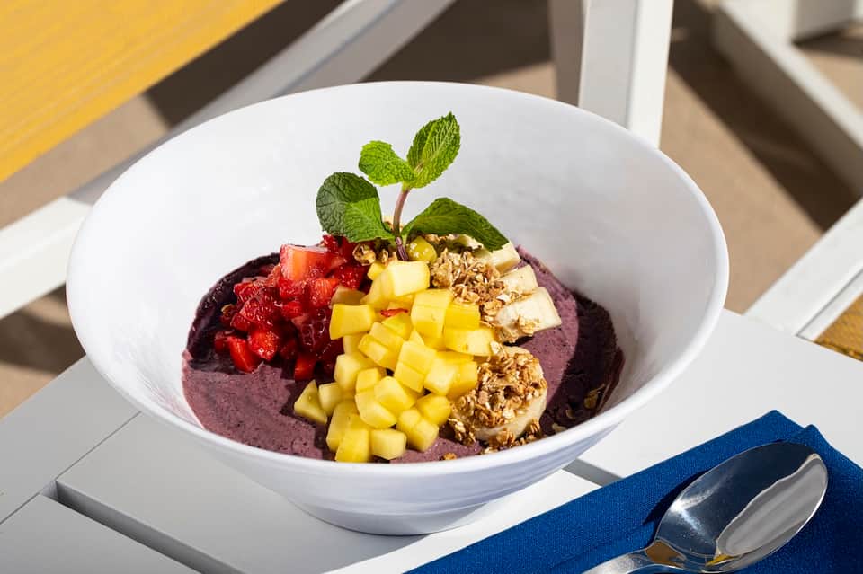 Gourmet bowl with purple base, fresh berries, yellow fruit chunks, granola, and mint leaf garnish on white plate